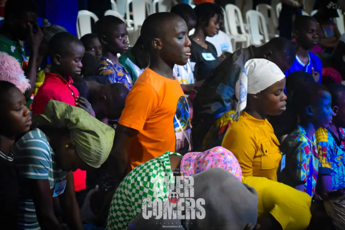 A group of children with heads bowed in prayer at an indoor camp event.